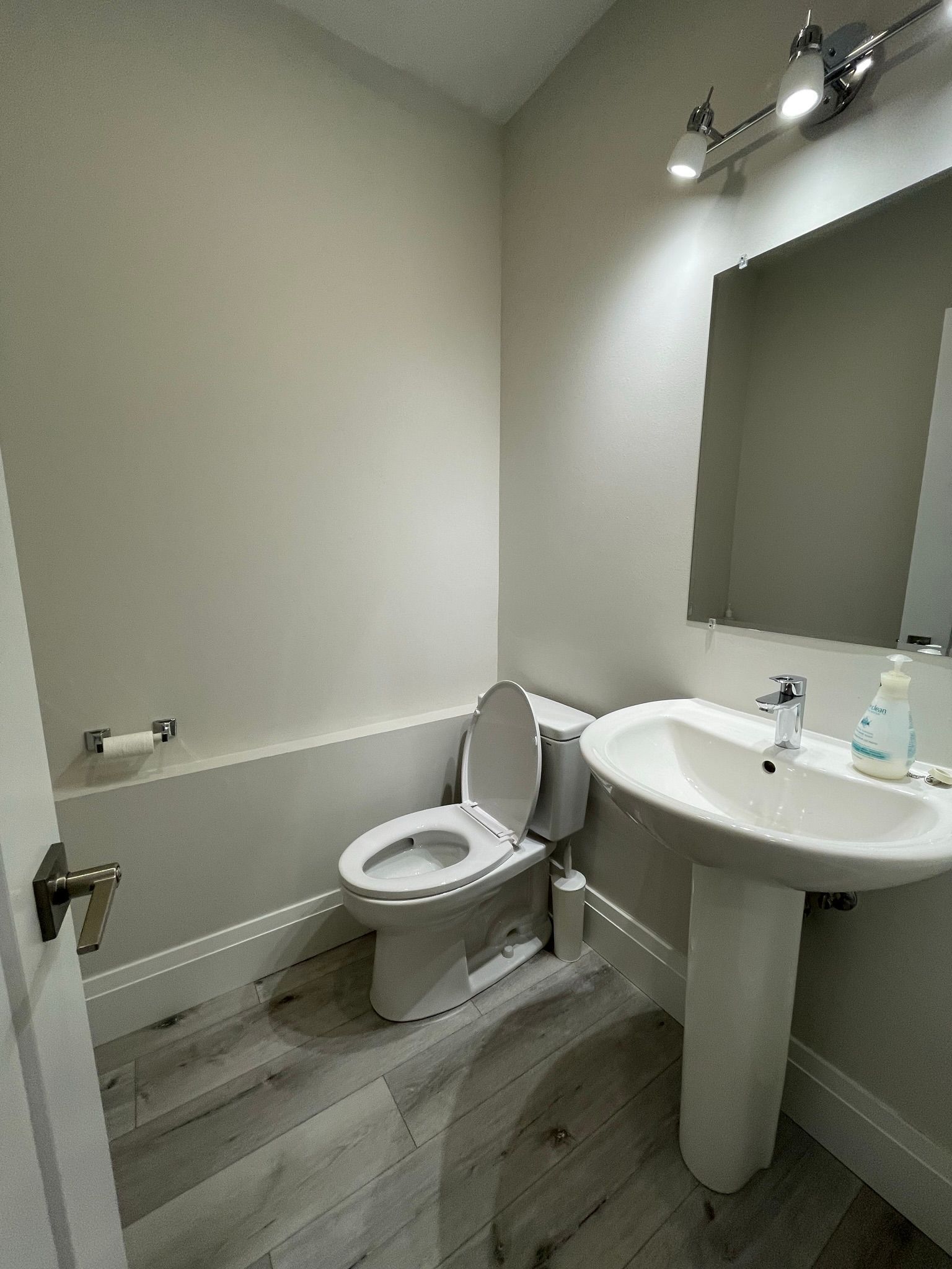 A small, neutral-toned bathroom with a toilet, pedestal sink, and mirror.  Wooden-look floor.