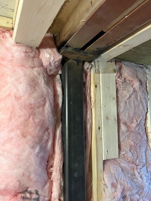 Pink insulation surrounds a black pipe in a wall, near wooden beams.