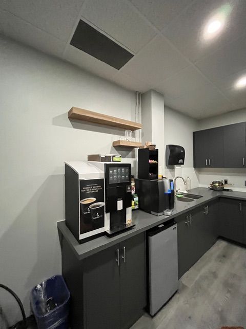 Office break room with coffee machines, sink, and cabinets; grey and white color scheme.