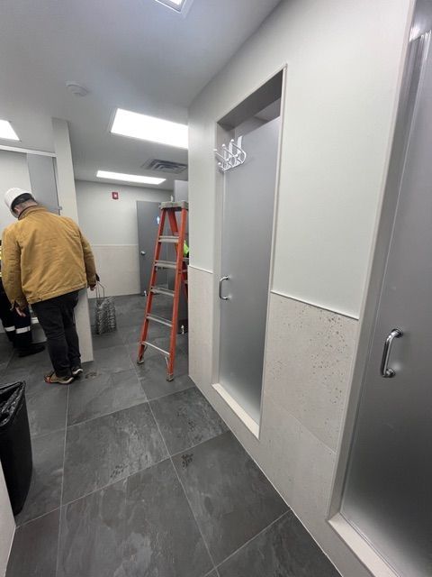 A bathroom with two frosted glass shower stalls and a person near a ladder; gray tile floor.