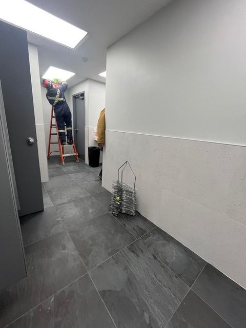 Person on ladder working on ceiling in a narrow hallway with gray tile flooring and white walls.
