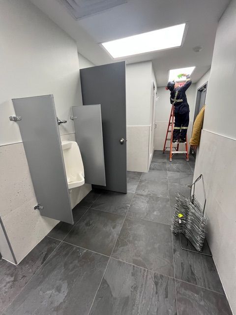 Public restroom with gray tile floor, urinals, and a worker on a ladder near a skylight.