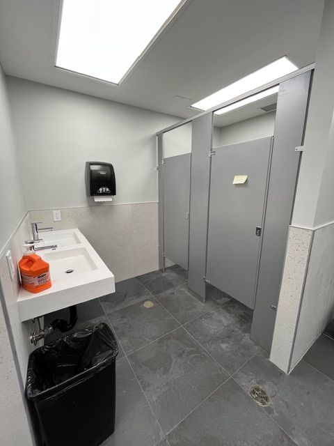 Public restroom with gray stalls, white sink, black trash can, and black tile floor.