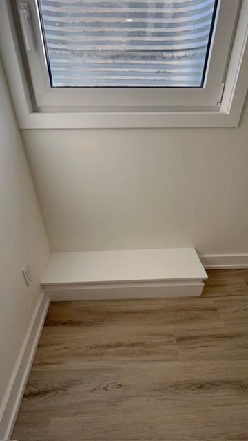 A white shelf built under a window, against a white wall and wood-look flooring.