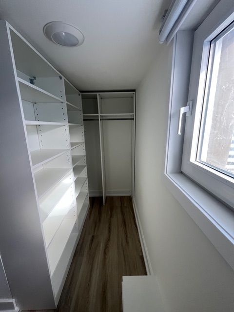 A narrow white walk-in closet with shelves and a clothing rod, wood-look floor, and a window.