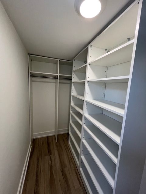 Walk-in closet with white shelves and rod. Wooden floor, neutral walls, and a ceiling light.