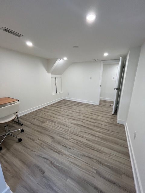 Empty white-walled room with wood-look flooring and recessed lighting. A desk and door are visible.