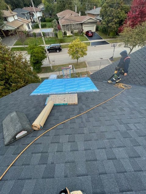 Roofer working on a dark gray shingled roof with safety equipment. A blue tarp covers a section. Suburban setting.