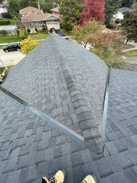 View of a newly shingled roof with dark grey shingles, metal flashing, and fall foliage in the background.