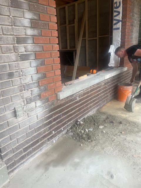 Man working on brick construction near a window opening, with mortar and an orange bucket present.