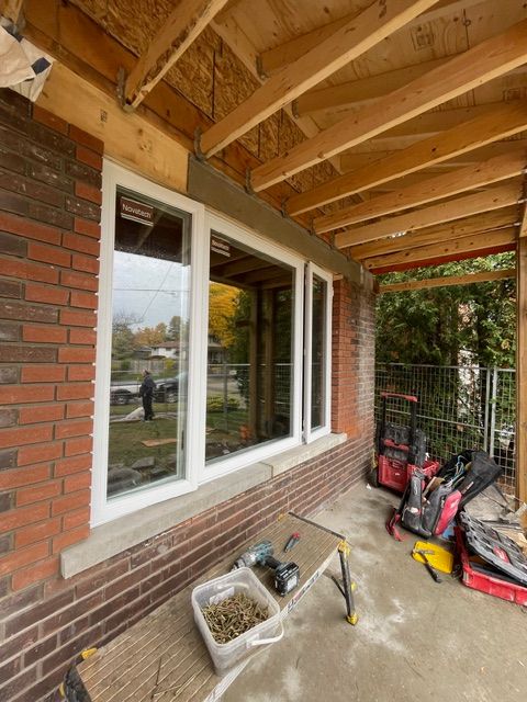 Exterior view of a brick house with new white-framed windows, under a wooden overhang. Construction tools visible.