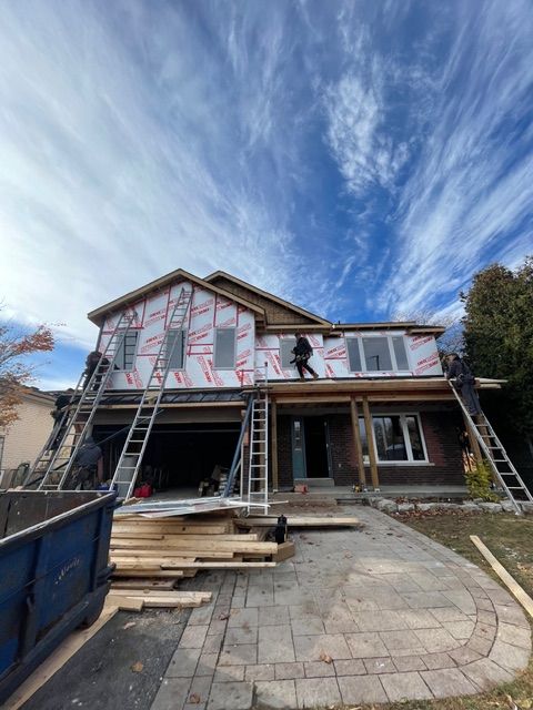 House under construction; workers on ladders applying siding. Blue sky with clouds. Wooden beams and materials on the driveway.