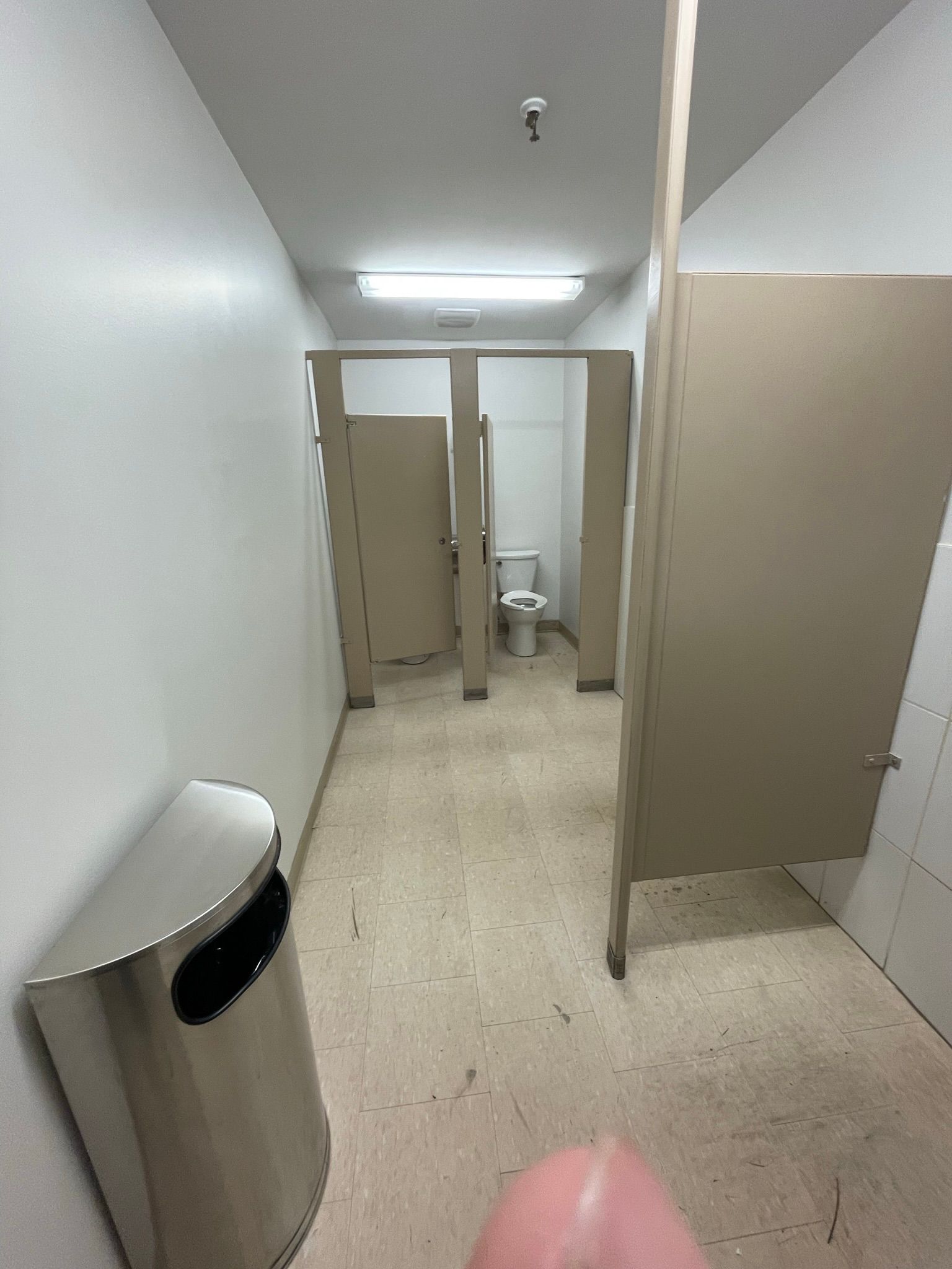 Public restroom with beige stalls and silver trash can. White walls and fluorescent lighting.