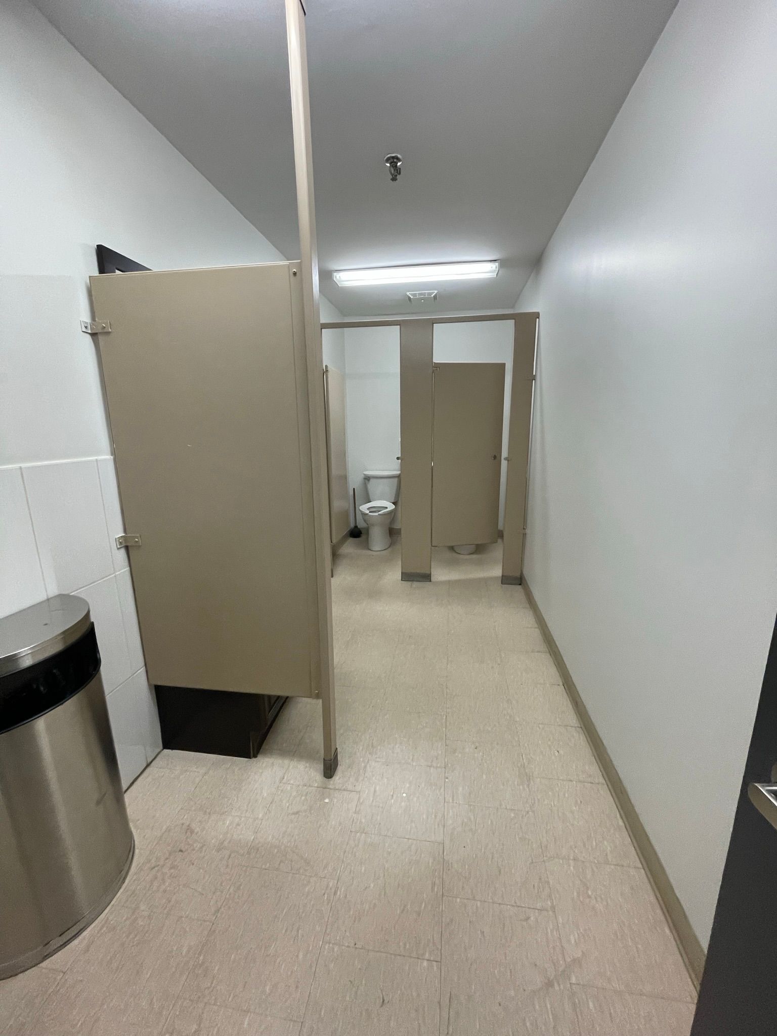 Narrow restroom hallway with stalls, a toilet, and a trash can.
