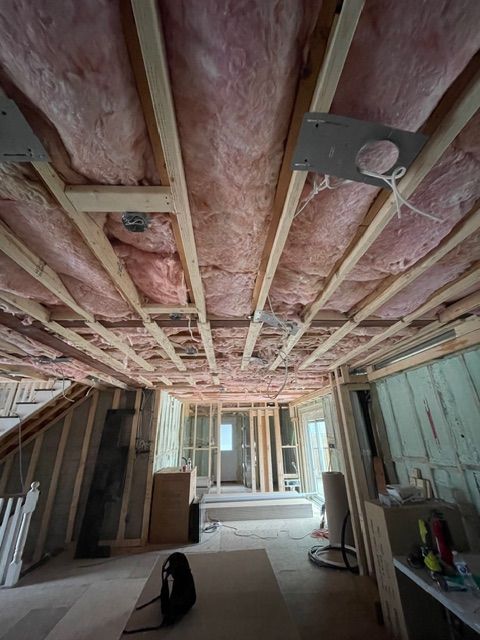 Interior view of a room under construction, showing exposed wooden beams, pink insulation, and partially framed walls.