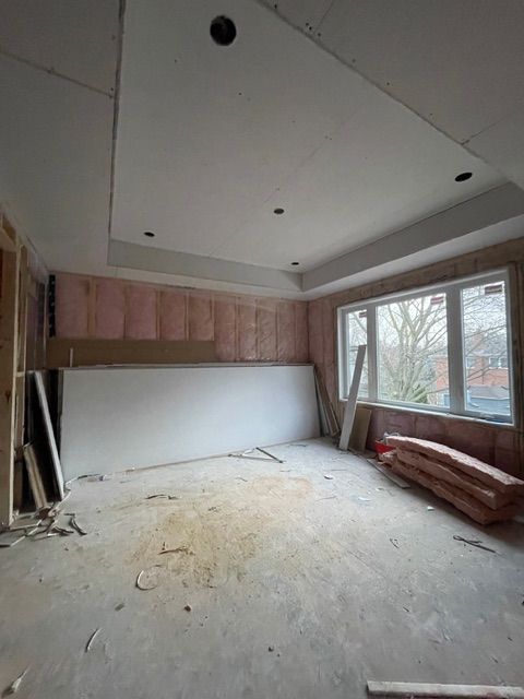 Room under construction, pink insulation, drywall leaning against wall, bay window, unfinished ceiling with recessed lighting.