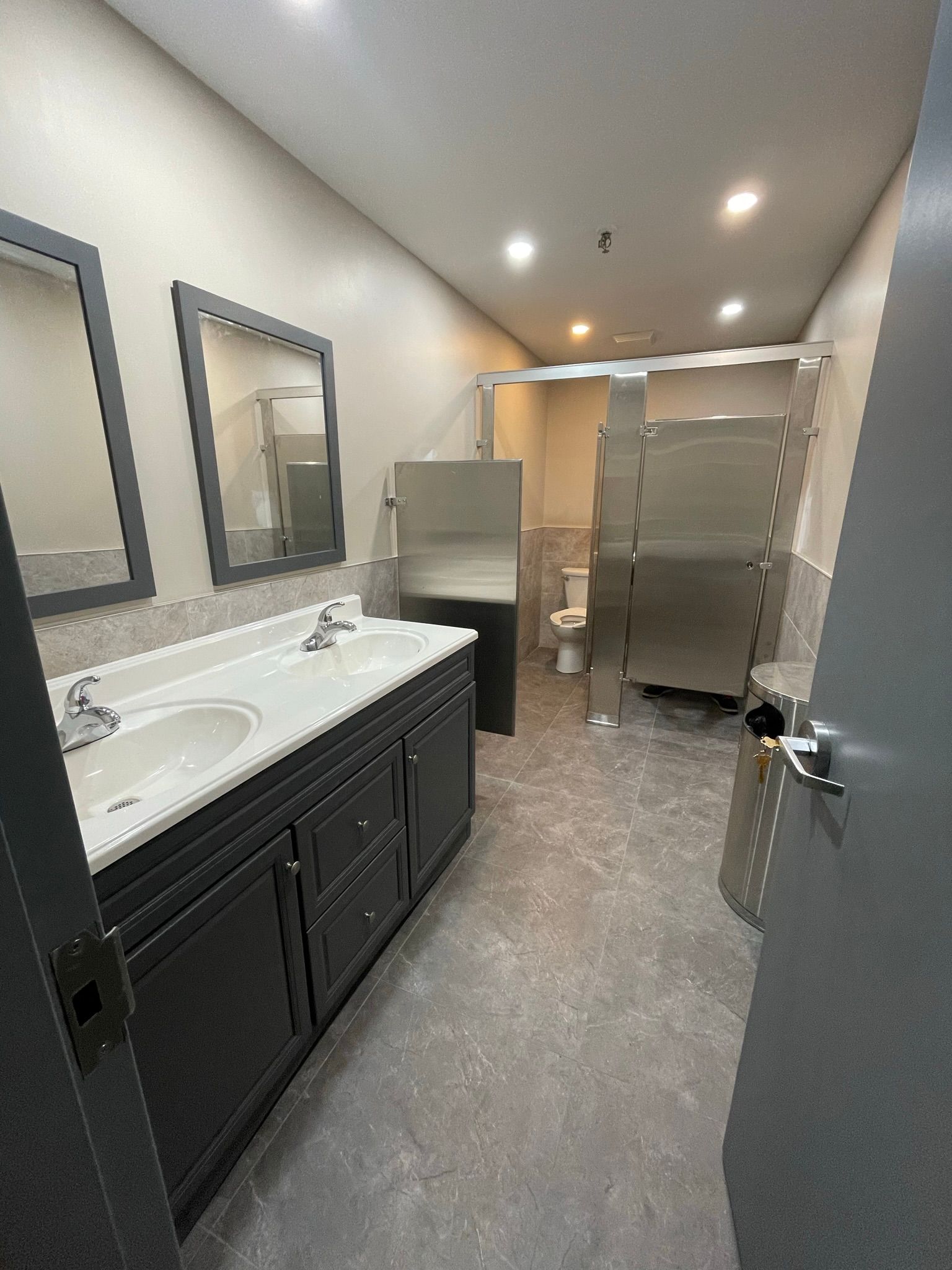 Public restroom with dual sinks, mirrors, and stalls, gray and stainless steel fixtures.