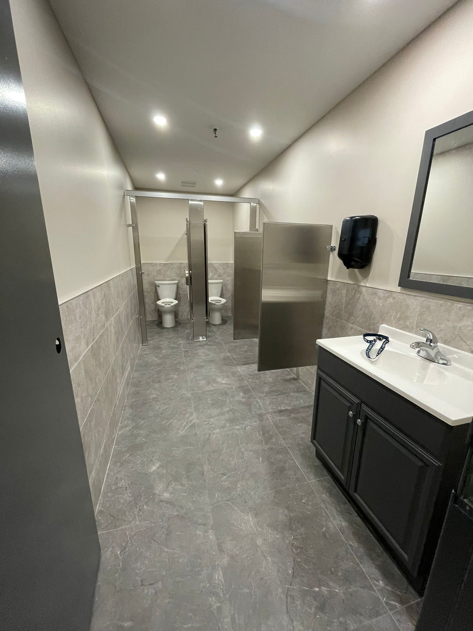 Public restroom with stalls, sinks, and mirrors. Gray and white color scheme.