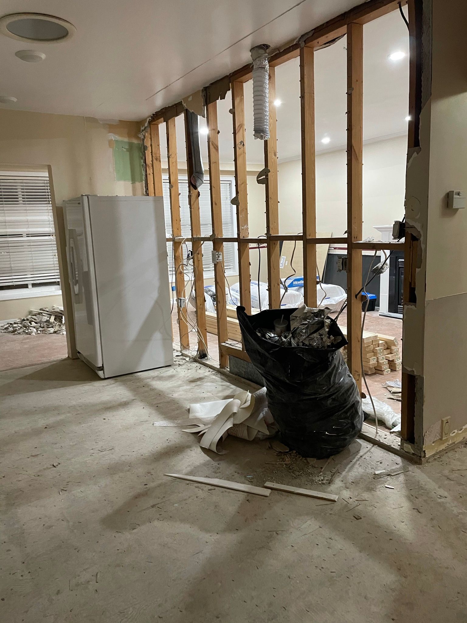 Interior demolition with exposed wooden wall studs, white refrigerator, trash bag, and debris on the floor.