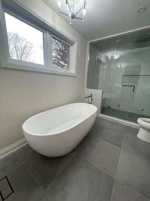 Bathroom with white oval tub, gray tile floor, glass shower, and window.