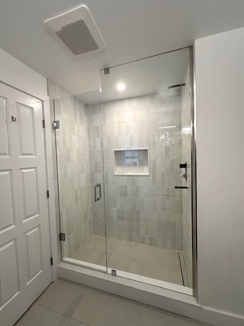 Modern shower with glass door, tile walls, recessed shelf, and white door to the left.