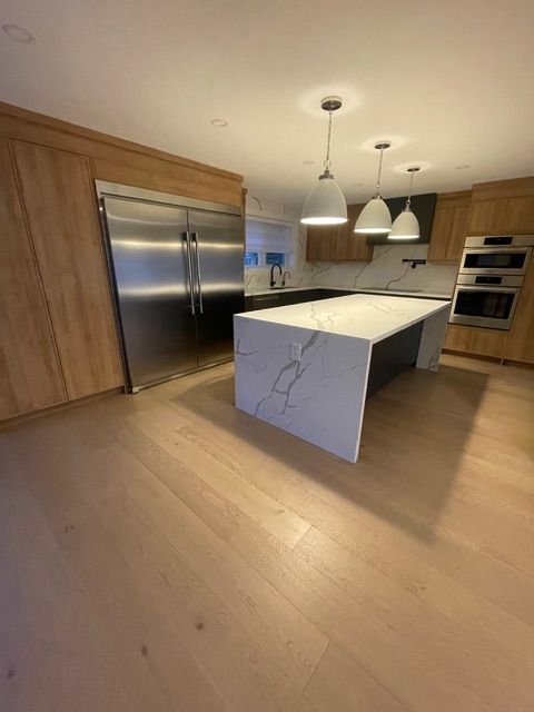 Modern kitchen with a large island, stainless steel refrigerator, light wood cabinets, and three pendant lights.