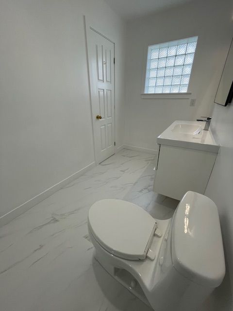 White bathroom with toilet, vanity, door, glass block window, and marble tile floor.