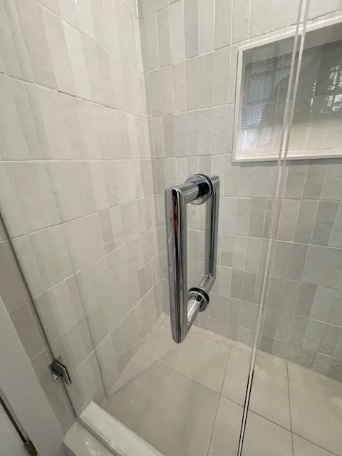 A shower with clear glass door, chrome handle, and white tiled walls.