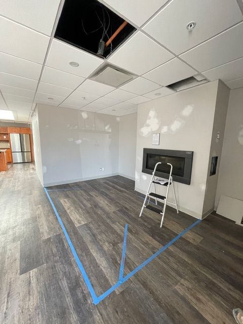 Room with wood-look floor, fireplace, and exposed ceiling panels. Blue tape outlines a section of the floor.