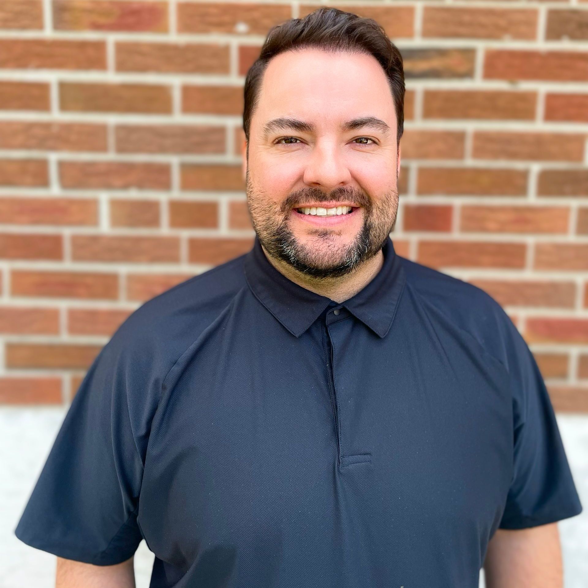 A man with a beard wearing a black shirt is smiling in front of a brick wall.