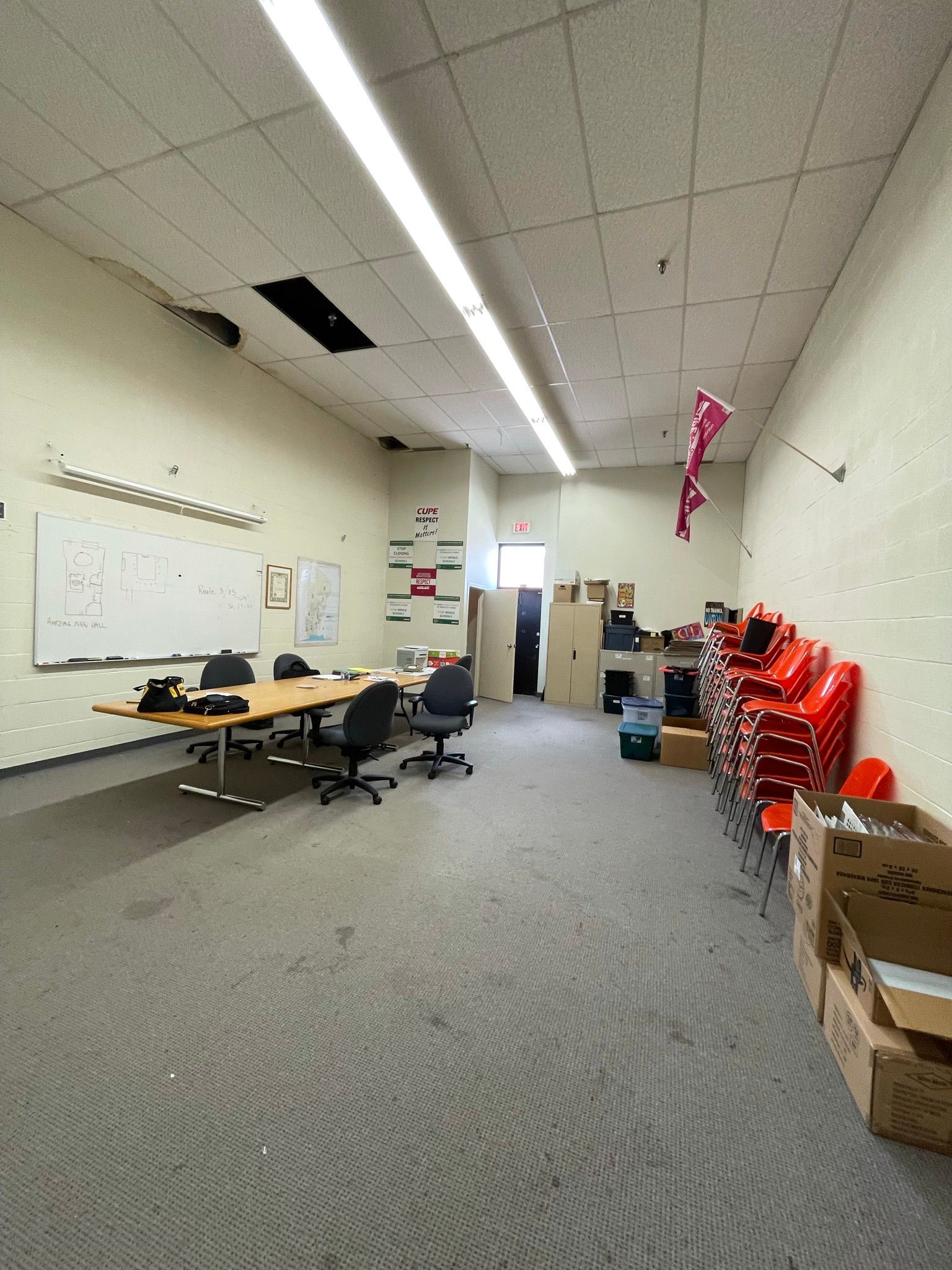 Empty office room with a long table, chairs, whiteboard, and various boxes and supplies.