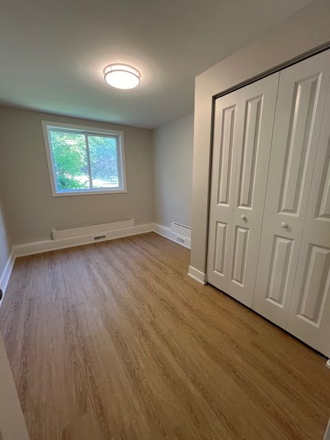 Empty bedroom with wood-look flooring, white closet doors, window, and neutral-colored walls.