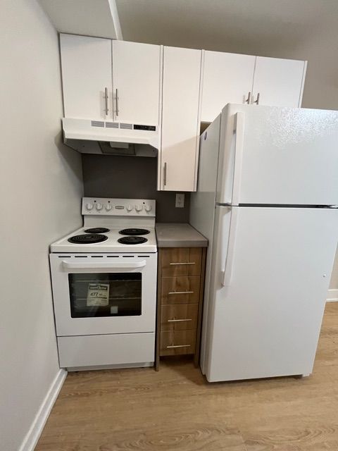 Small, white kitchen with oven, cabinets, refrigerator, and wooden cabinet, with wood flooring.