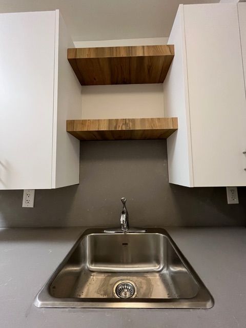 Stainless steel sink with faucet, gray backsplash, and wooden shelves between white cabinets.