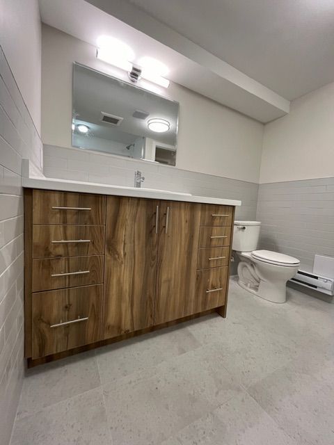 Bathroom with a wooden vanity, white countertop, and gray tiled floor and walls. A toilet sits to the right.