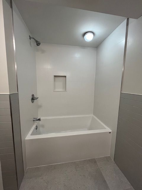 White-walled bathroom with a bathtub, showerhead, and recessed shelf. Grey floor and adjacent wall.