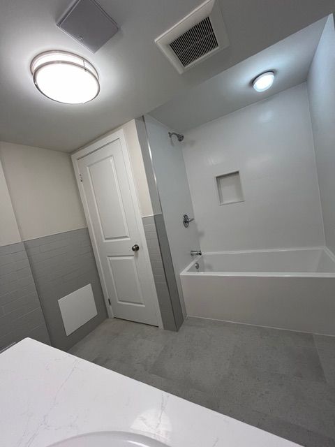 Bathroom with white and gray walls, white bathtub, door, and sink. Overhead lights.