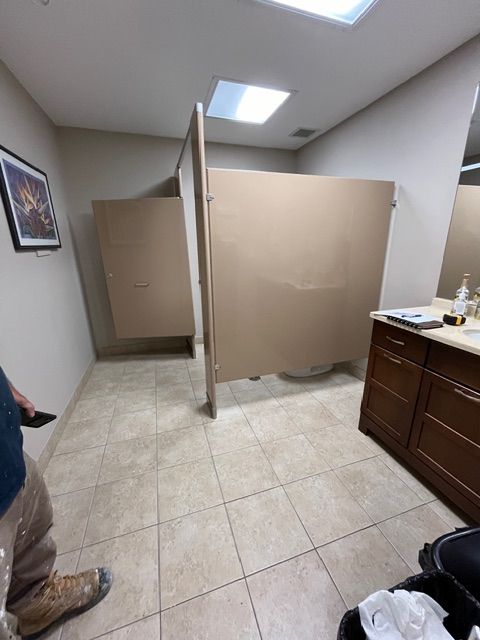 Public restroom with two stalls, beige doors, tile floor, vanity, and a person.