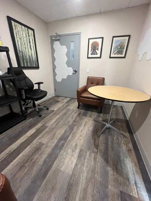 Office with dark wood-look floor, black chair, round table, brown armchair, door with honeycomb cutouts, and artwork.