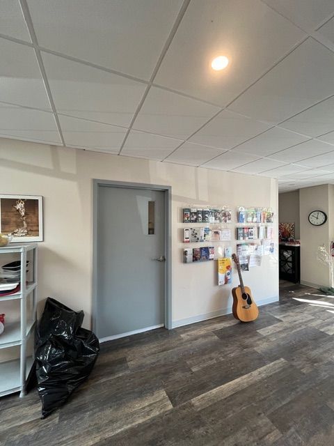 A gray door and guitar on a wood-look floor; a bulletin board and a black trash bag are nearby.