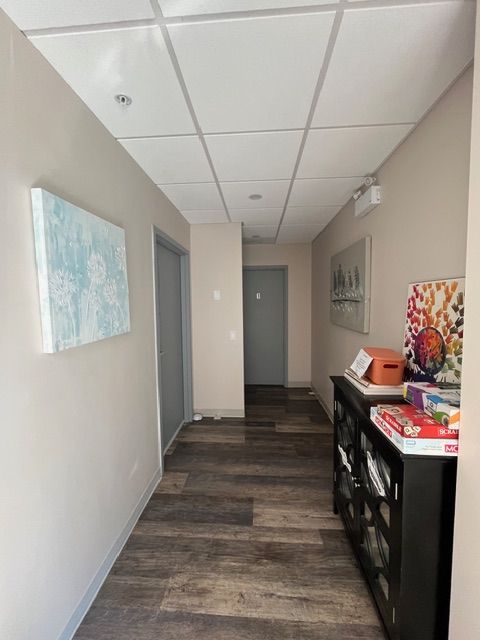 A narrow hallway with tan walls, wood-look flooring, and a drop ceiling.  A few closed doors, artwork, and a shelf.