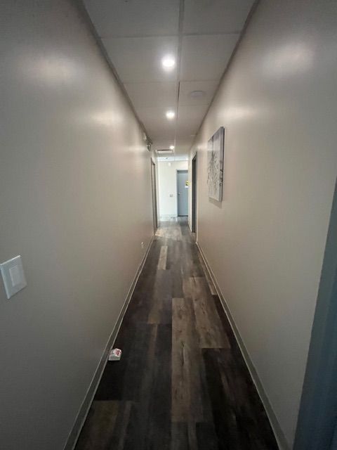 Narrow hallway with gray walls, wood-look floor, and overhead lights leading to a doorway.