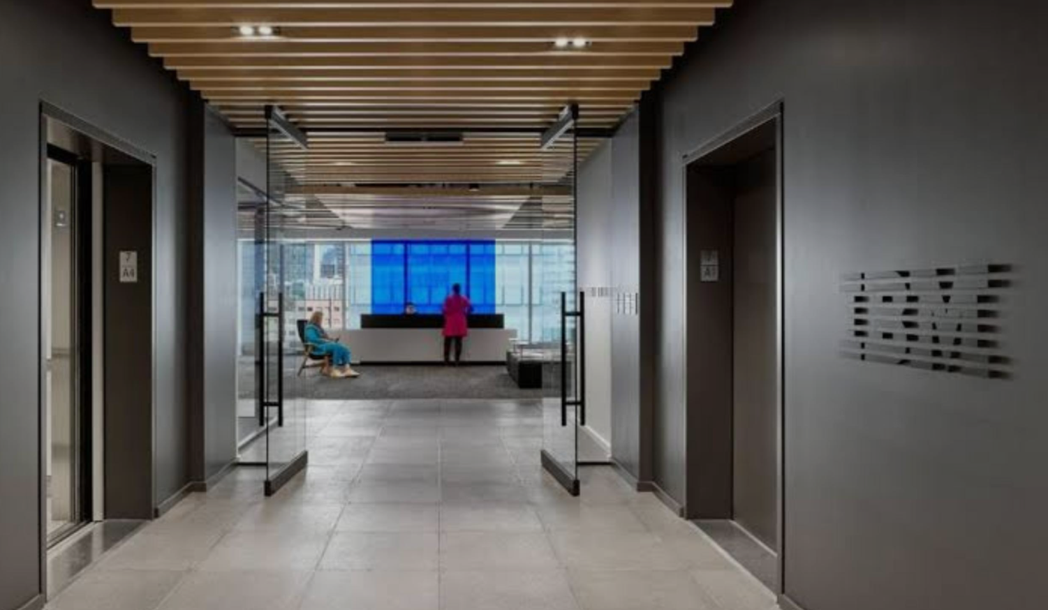 Hallway with glass doors and elevators. Person in pink coat near large blue window.