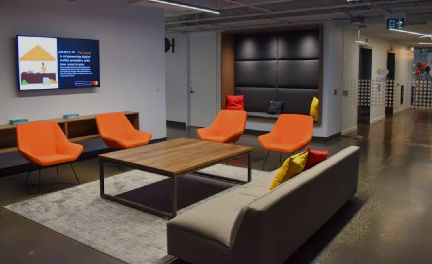 Lounge area with orange chairs, a gray sofa, wooden coffee table, and a screen displaying information, in a modern building.