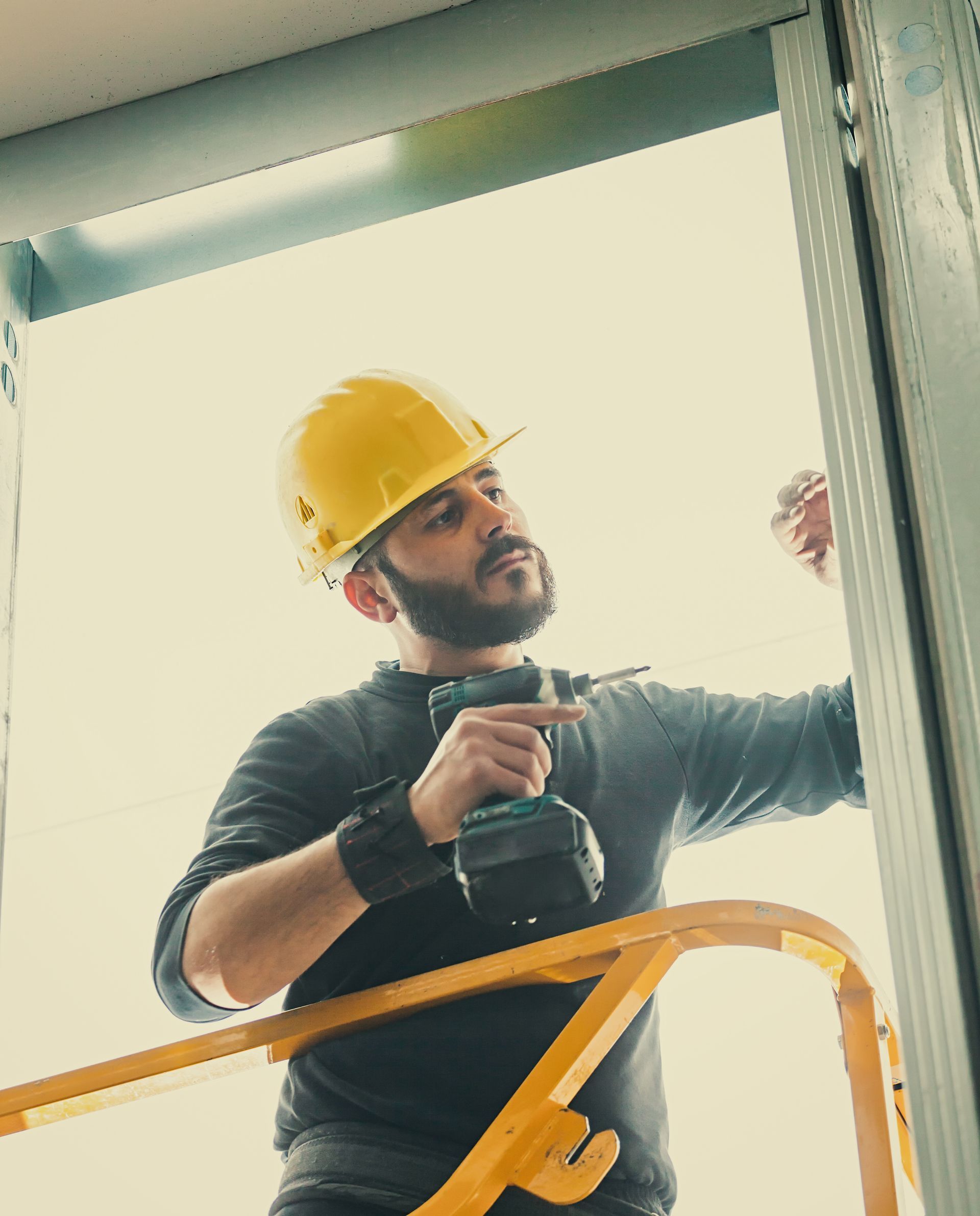 A man wearing a hard hat is using a drill on a ladder.