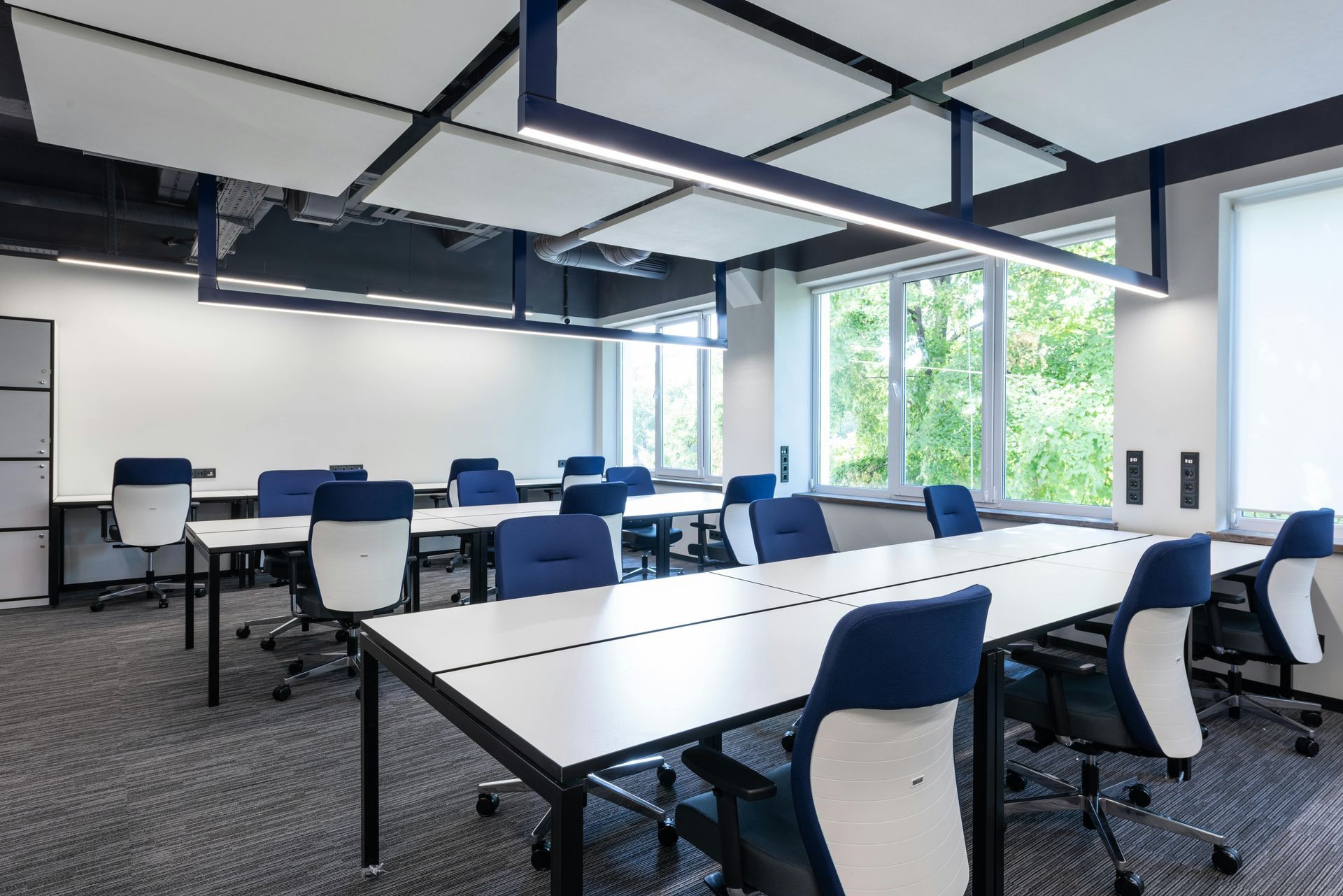 Office space with white desks, blue chairs, and a window with a view of greenery.