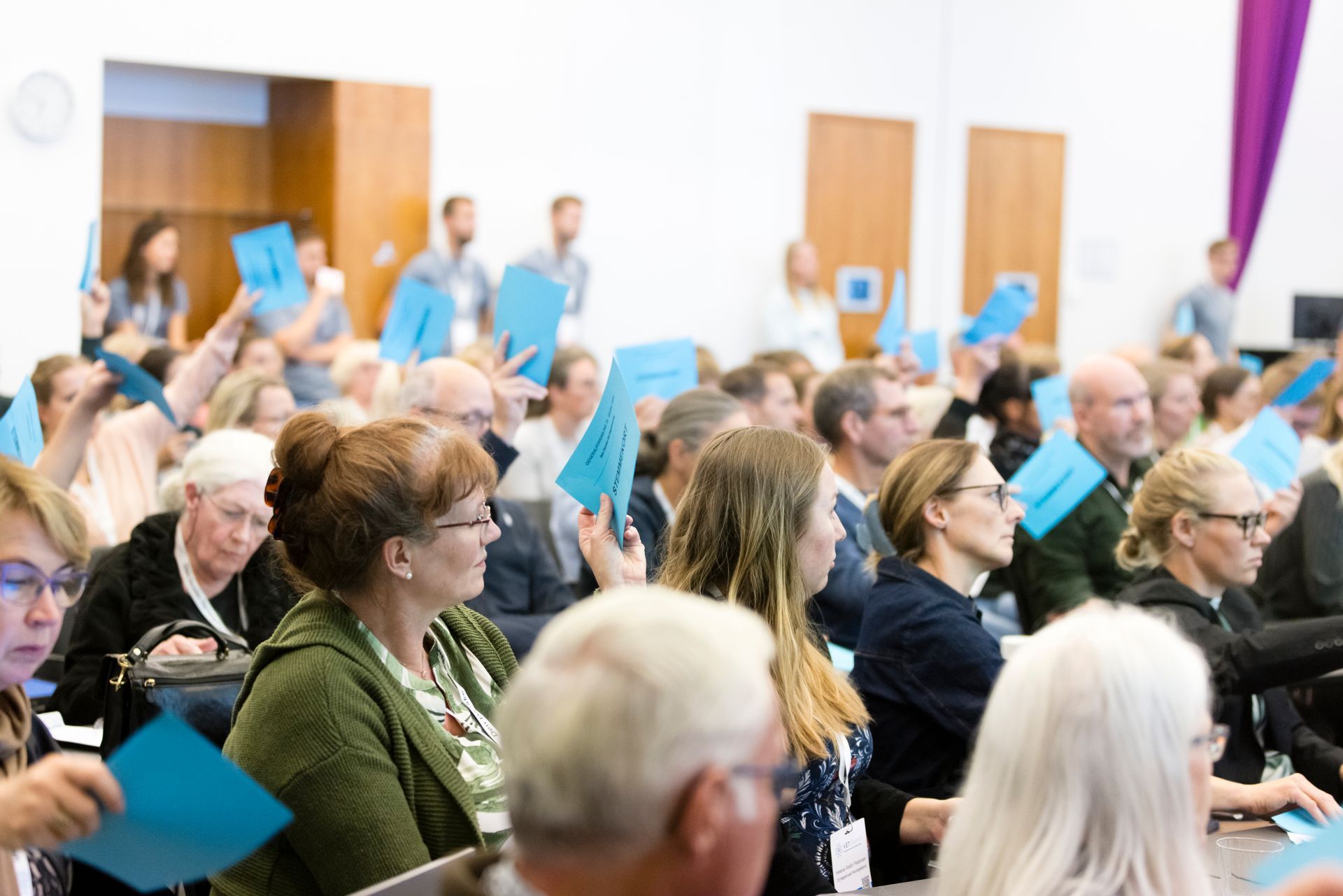 Audience in a large room raise blue cards, likely at a conference or event.