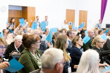 Audience in a large room raise blue cards, likely at a conference or event.