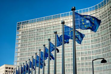 European Union flags wave in front of a modern, glass-walled building in bright sunlight.