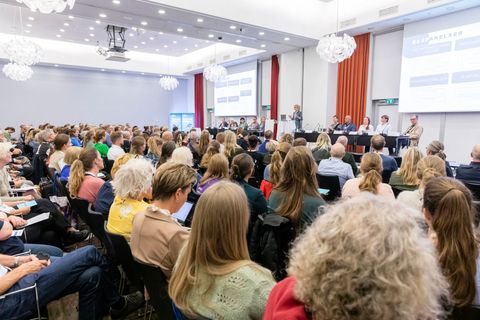 Large conference hall filled with people attending a presentation; a panel of speakers is at the front.
